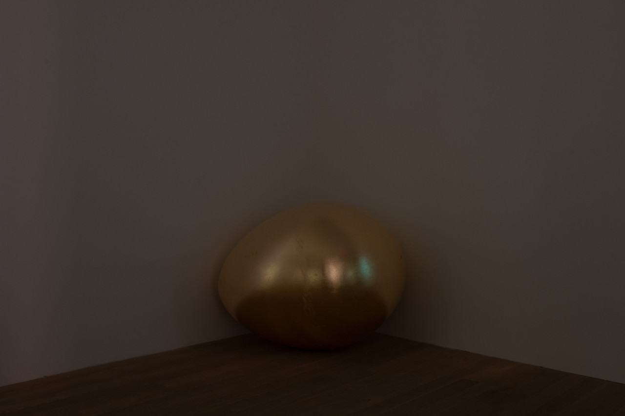 Color image of a corner in a darkened gallery space. A large golden egg sculpture sits on the floor in the corner.