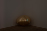 Color image of a corner in a darkened gallery space. A large golden egg sculpture sits on the floor in the corner.