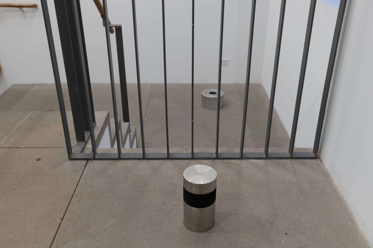 Color image of two small doorstop sculptures on the floor of the gallery space landing. The sculpture in the foreground consists of a short column of aluminum, banded with black rubber near the top. In the background sits another sculpture, a squat column of concentric stainless steel rings with a black rubber core.