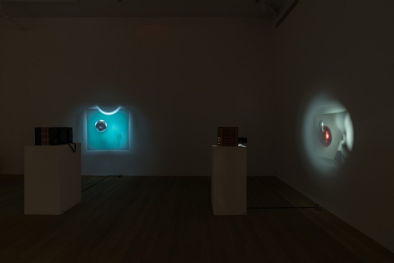 Color image of the corner of a darkened gallery installation. On the left, a pinhole projector inside a plaid travel bag sits atop a pedestal. On the back wall, an image of a green square with a crenellated metal opening is projected. On the right, a pinhole projector housed within a plywood box sits on a pedestal. On the right wall, an image of a red pearlescent ball, with a small white pearlescant ball attached on top, is projected.