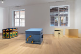Color image of a gallery installation containing four large wooden box sculptures by Matthew Langan-Peck, three of which are slightly open and have headphones hanging from their sides. To the left, a dark blue box is painted with a repeated traffic light motif. The box in the center foreground is messily painted sky blue, with two yellow shooting stars on one side. In the background, to the right, two wooden boxes sit next to each other, one painted light yellow and the other covered in imitation gold leaf.