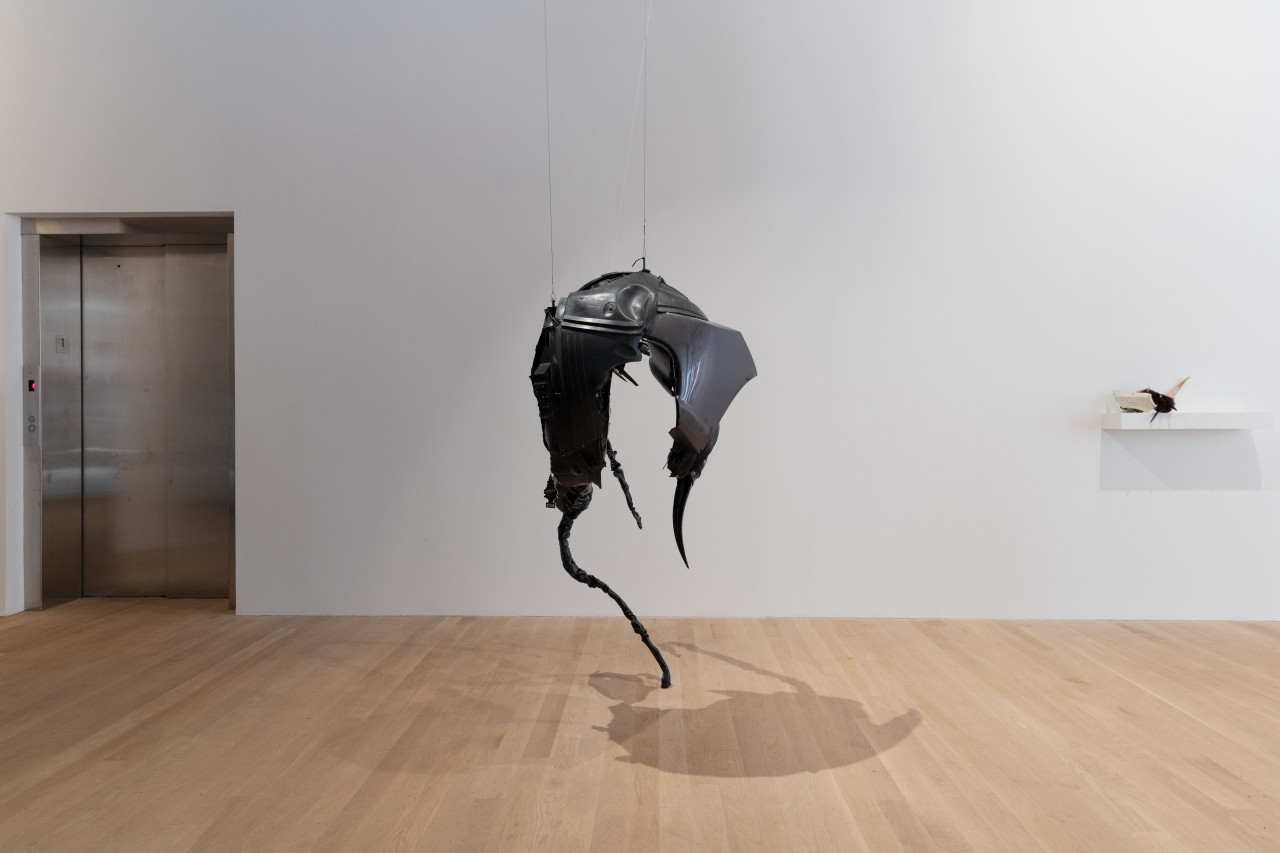 A hulking biomorphic black sculpture, composed of car parts and a large black t-shirt smeared with pigmented dragon skin silicone, hangs from the ceiling. To the right, a small sculpture of scuffed white plastic, brown fur, and various jutting animal teeth sits on a shelf.