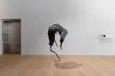 A hulking biomorphic black sculpture, composed of car parts and a large black t-shirt smeared with pigmented dragon skin silicone, hangs from the ceiling. To the right, a small sculpture of scuffed white plastic, brown fur, and various jutting animal teeth sits on a shelf.