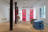 Image of a gallery space installed with multiple sculptures. On the right, two large painted wooden boxes by Matthew Langan-Peck are slightly open and have headphones hanging on their sides. The boxes are painted light blue with yellow shooting stars and dark blue with traffic light-esque designs, respectively. On the left, two of Ruben Ulises Rodriguez Montoya's large biomorphic sculptures, composed of found materials, hang from the ceiling. In the background, Isabelle Frances McGuire's three large pink fabric banners emblazoned with silver stars hang from ceiling to floor.