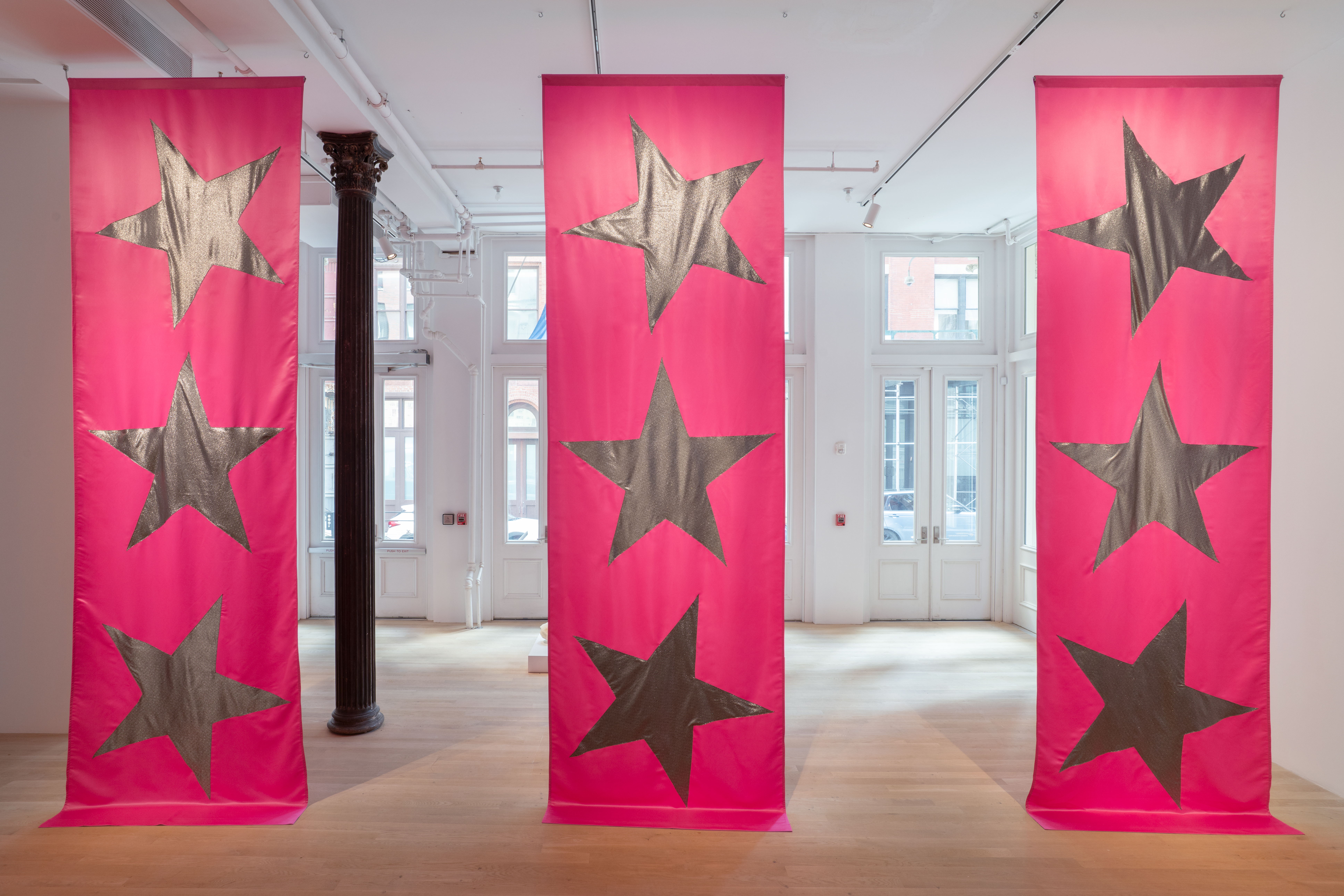 Three large pink fabric banners by Isabelle Frances McGuire, each bearing three silver star patches, hang from the ceiling to rest on the floor.