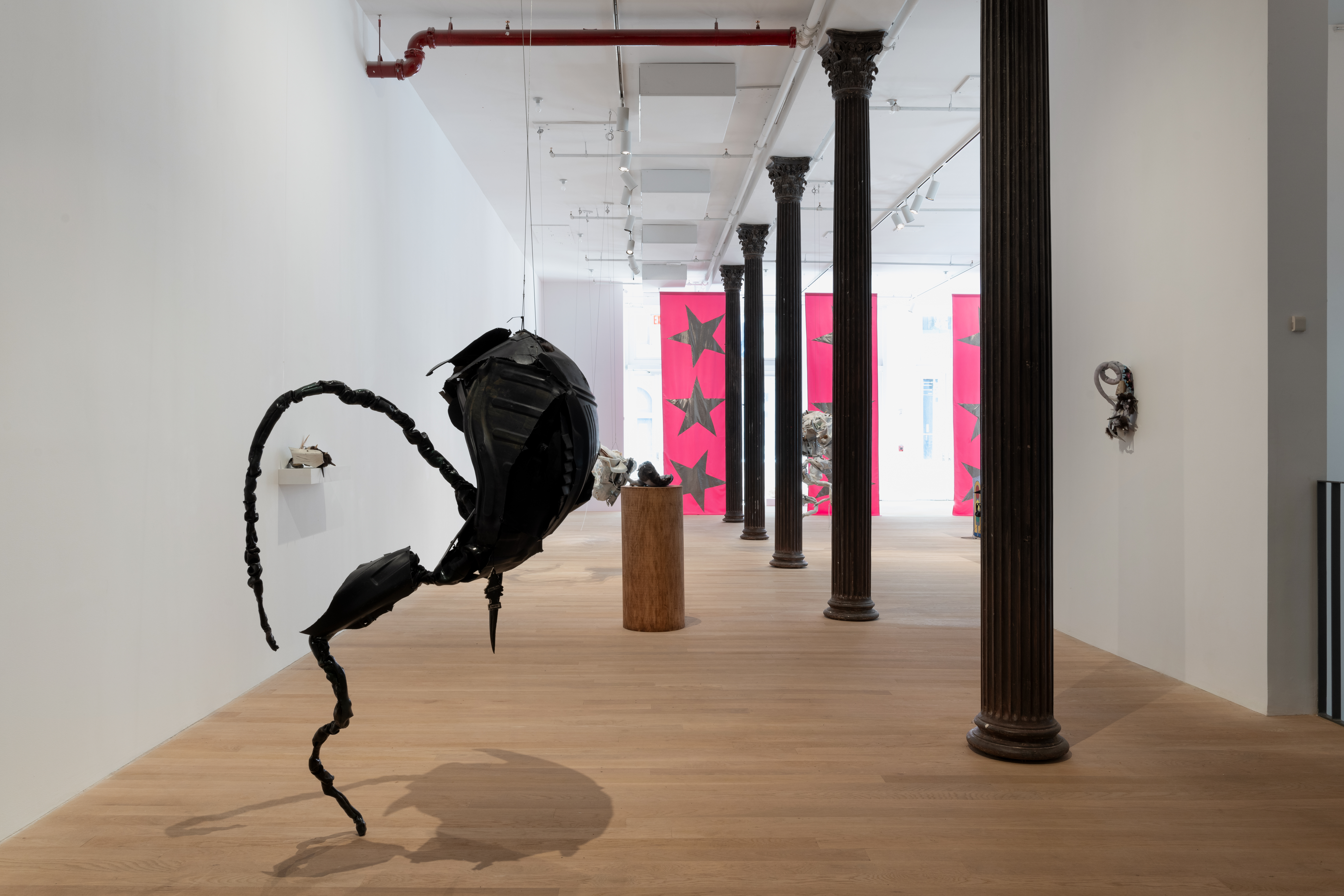 A large black biomorphic sculpture hangs in the foreground. In the distance, various biomorphic sculptures hang from the ceiling, sit on pedestals, or hang from the walls, receding deeper into the gallery. Three large pink banners emblazoned with silver stars hang from the ceiling in the background.