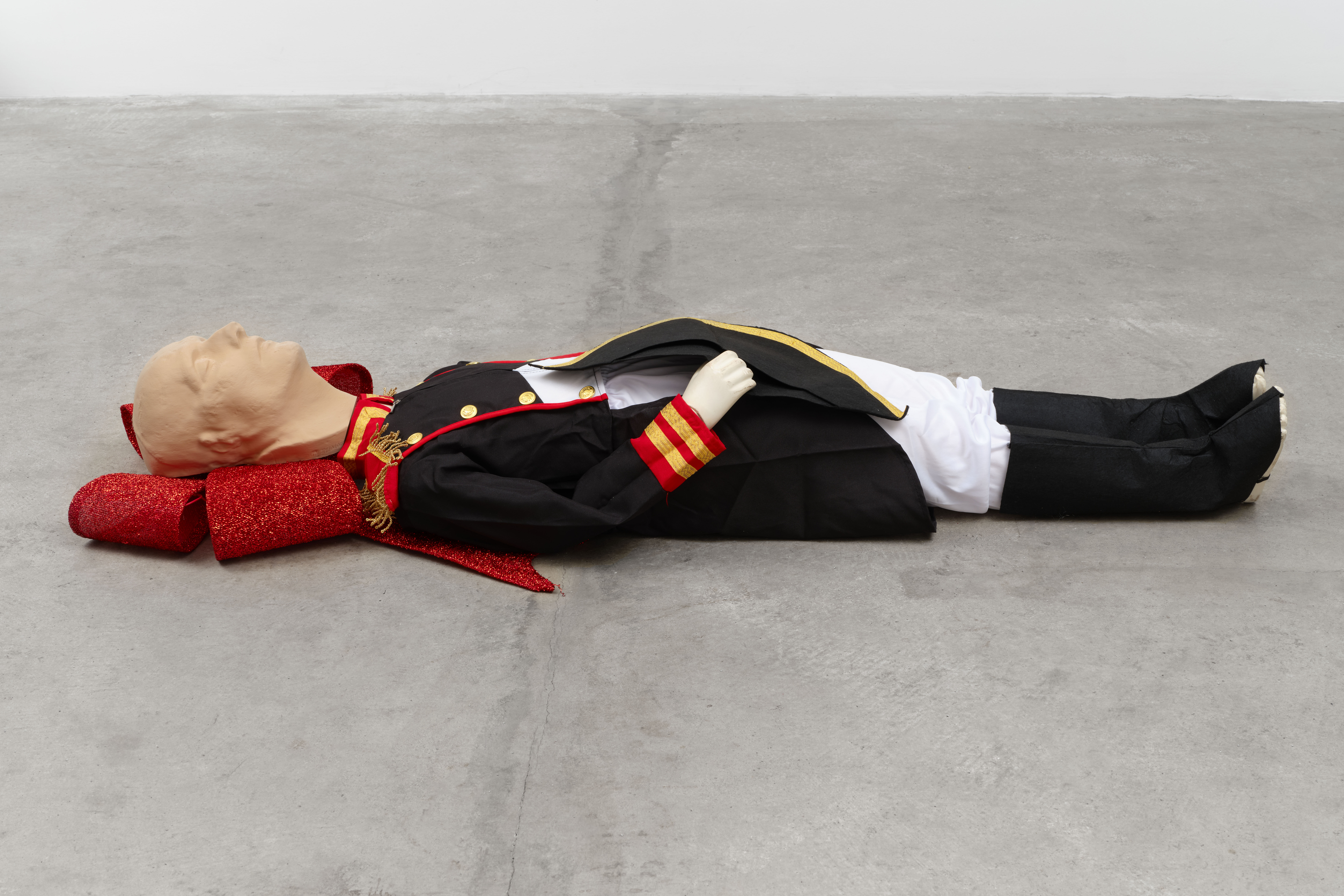 Color image of a sculpture of a human body, laying face up, placed on a concrete floor. The figure is dressed in French military apparel and their head rests on a large glittery, red bow.
