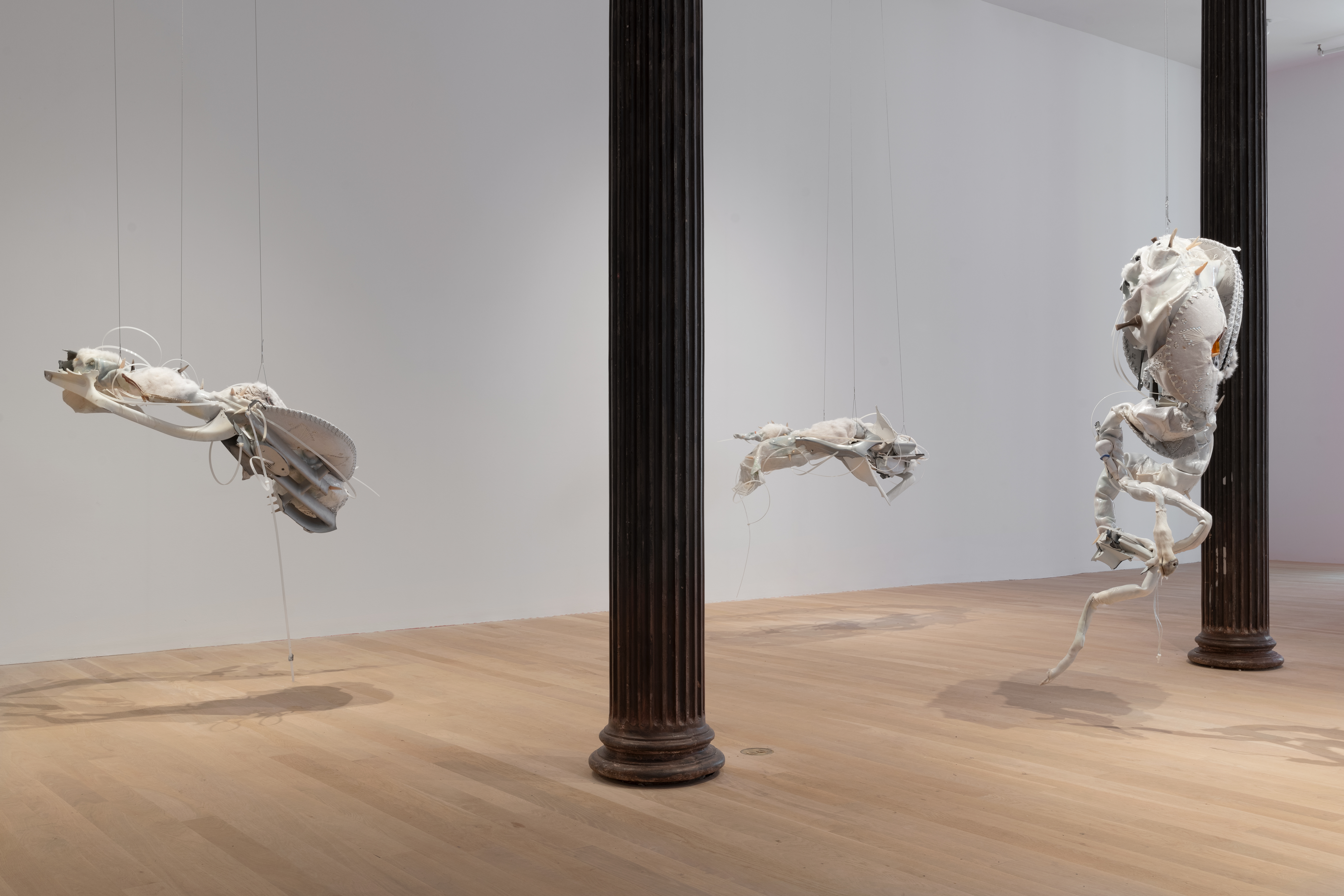 Color image of a gallery space with three large white biomorphic sculptures by Ruben Ulises Rodriguez Montoya hanging from the ceiling throughout. The sculptures are composed of discarded car parts, pigmented silicone, animal horns, clothes, and various other found materials.