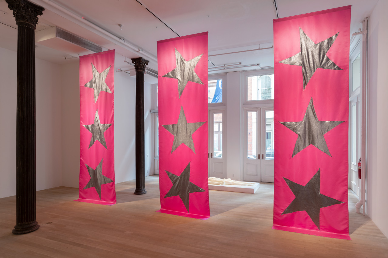 Color image of three large pink fabric banners by Isabelle Frances McGuire, each emblazoned with three silver stars, hanging from ceiling to floor. In the gallery space behind the banners, an off-white foam sculpture of a naked human form rests supine on a small white platform.