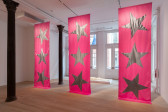 Color image of three large pink fabric banners by Isabelle Frances McGuire, each emblazoned with three silver stars, hanging from ceiling to floor. In the gallery space behind the banners, an off-white foam sculpture of a naked human form rests supine on a small white platform.