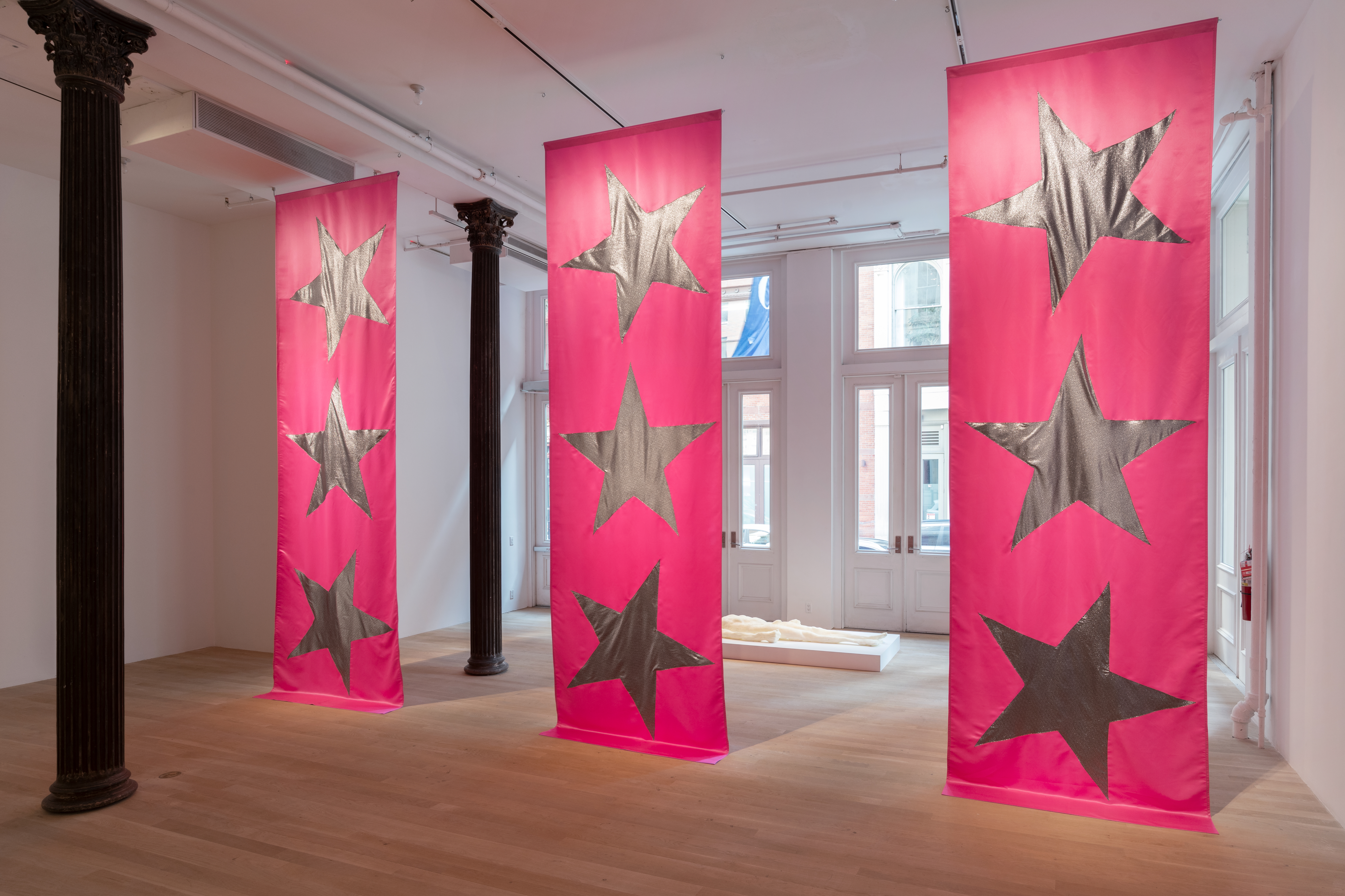 Color image of three large pink fabric banners by Isabelle Frances McGuire, each emblazoned with three silver stars, hanging from ceiling to floor. In the gallery space behind the banners, an off-white foam sculpture of a naked human form rests supine on a small white platform.