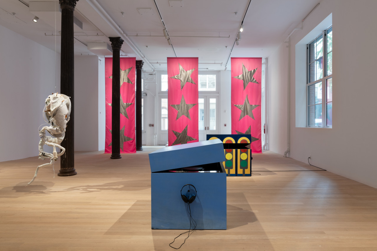 A view of a gallery installed with multiple sculptures by Matthew Langan-Peck, Ruben Ulises Rodriguez Montoya, and Isabelle Frances McGuire. In the center foreground sits a wooden box sculpture painted light blue, with a pair of headphones hanging from a small peg on the side facing us. Another wooden box, painted dark blue with a repeated traffic light motif, sits in the distance behind and partially occluded by the box in the foreground. To the left, a large biomorphic sculpture composed of found materials hangs from the ceiling. In the background, three large pink banners emblazoned with silver stars hang from ceiling to floor.