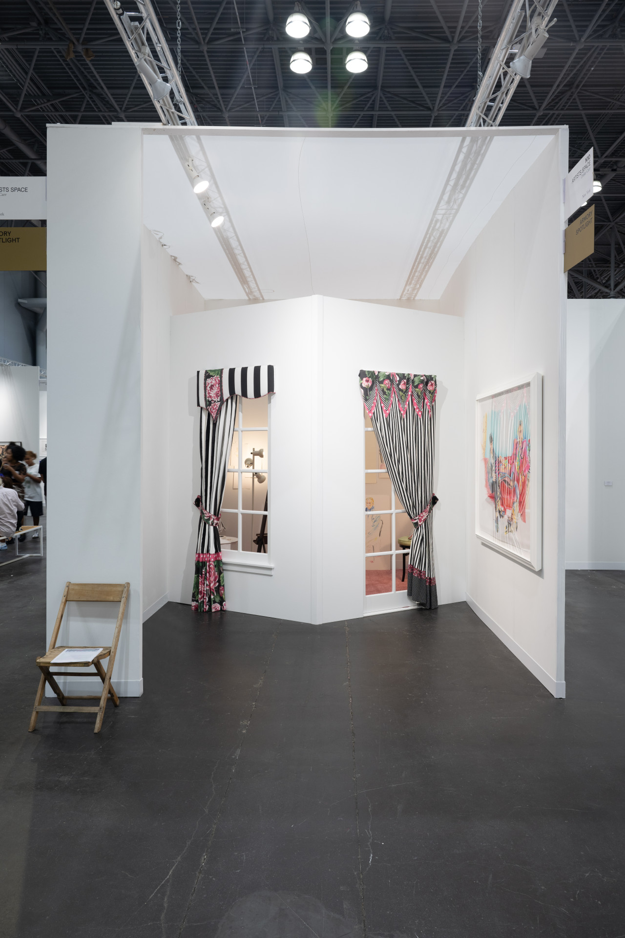 Color image of front view of a booth inside an art fair. On the right wall hangs a colorful drawing.  At the center of the booth, an installation shows the corner of a domestic space, with a window on each side and black and white curtains with pink flowers. Through the windows, the leg of a chair, a lamp, an easel, and some drawings can be seen.