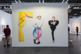 Color image of a booth at an art fair. At the center of the image, there is a white wall with three works made out of cut-out panel. The one on the left is cut in the shape of curtains, which are yellow with pink ruffles. The work in the center is a male-presenting figure with one hand above its head, and the other one in its pocket. It's wearing a white jacket, black shorts, pink tights, and black shoes. The work on the right is a woman-presenting figure, with short blond hair. It has its arms crossed over the chest, and is dramatically looking up. It's wearing a black suit and a single red glove.