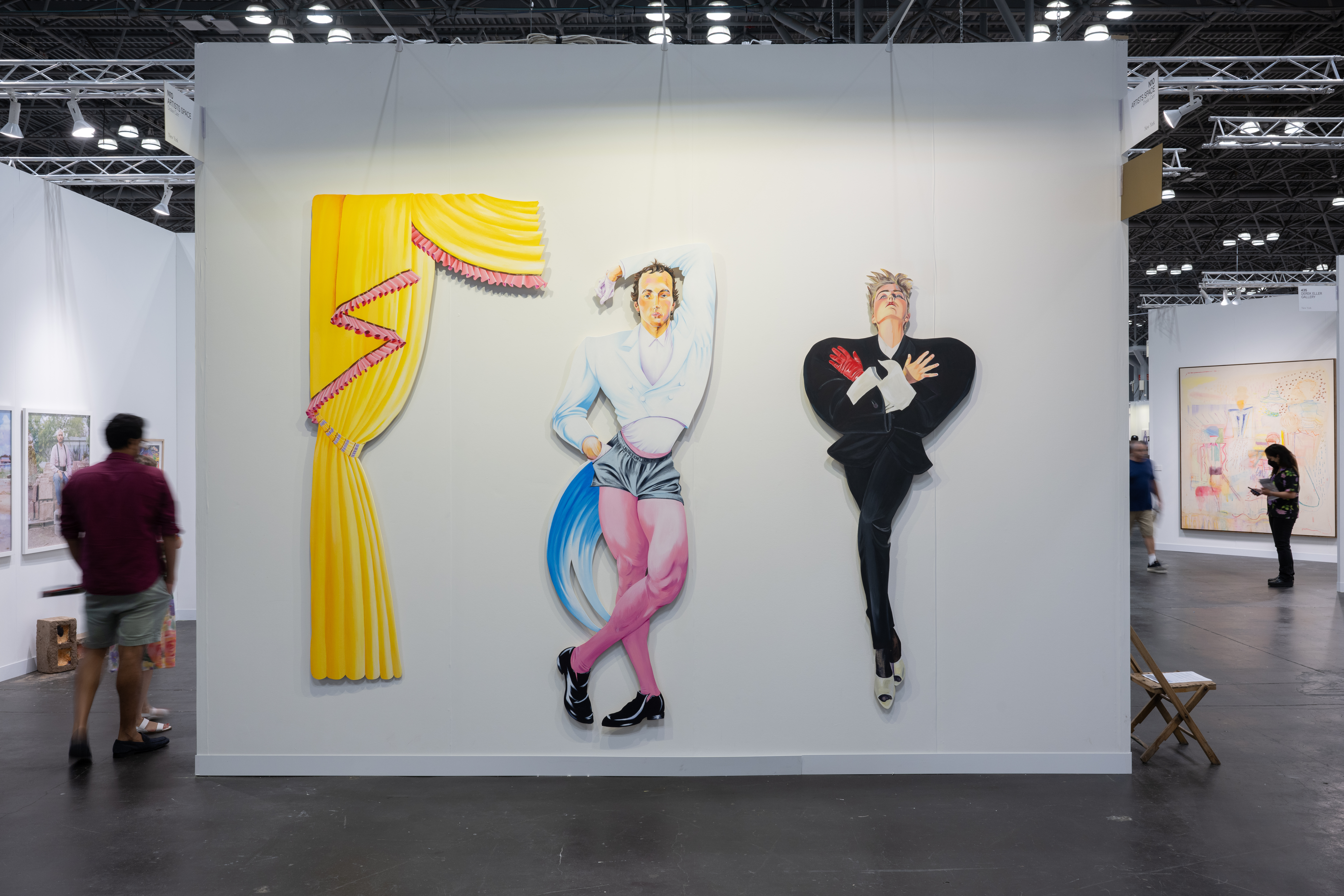 Color image of a booth at an art fair. At the center of the image, there is a white wall with three works made out of cut-out panel. The one on the left is cut in the shape of curtains, which are yellow with pink ruffles. The work in the center is a male-presenting figure with one hand above its head, and the other one in its pocket. It's wearing a white jacket, black shorts, pink tights, and black shoes. The work on the right is a woman-presenting figure, with short blond hair. It has its arms crossed over the chest, and is dramatically looking up. It's wearing a black suit and a single red glove.