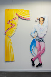 A color image of a white wall with two works made out of cut-out panel. The work on the left is cut in the shape of curtains, which are yellow with pink ruffles. The work on the right is a male-presenting figure posing with one hand above its head, and the other one in its pocket. It's wearing a white jacket, black shorts, pink tights, and black shoes.
