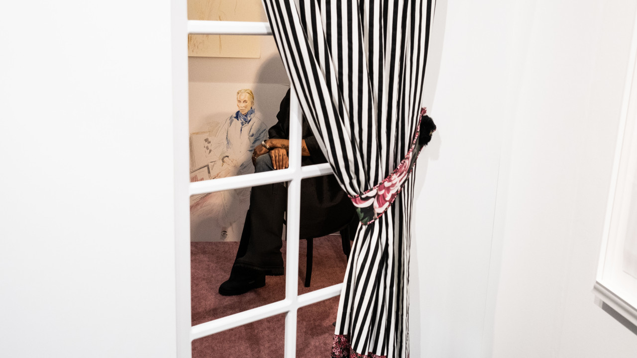 A color image of a white wall with windows and black and white curtains. Through the window, a cross-legged figure is partially seen sitting on a chair. Only the legs and hands are visible.