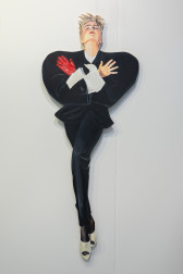 A color image of a work, made out of cut-out panel, of a woman-presenting figure with short blond hair. The work on the left is a woman-presenting figure, with short blond hair. It has its arms crossed over its chest, and is dramatically looking up. It's wearing a black suit and a single red glove