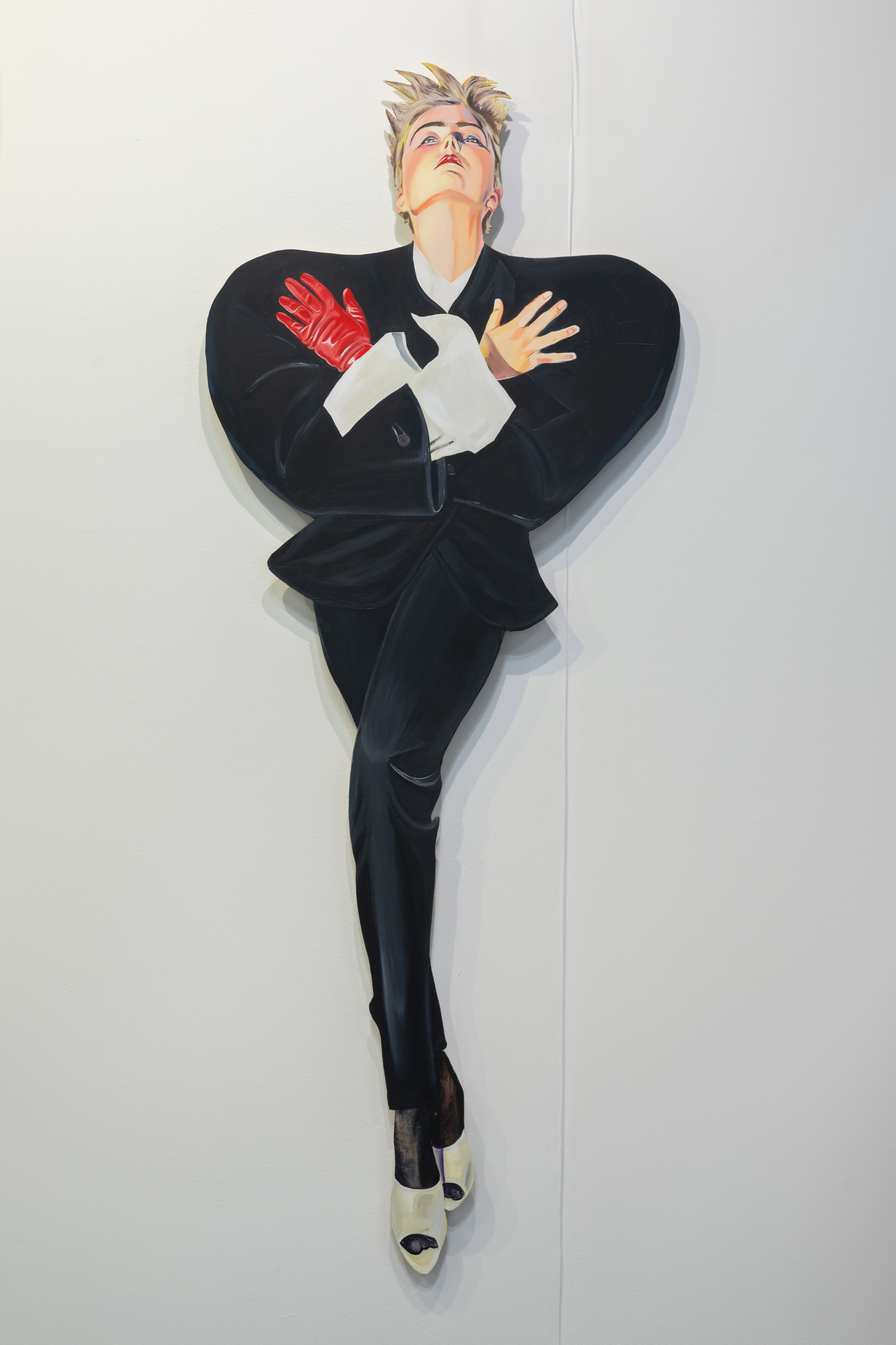 A color image of a work, made out of cut-out panel, of a woman-presenting figure with short blond hair. The work on the left is a woman-presenting figure, with short blond hair. It has its arms crossed over its chest, and is dramatically looking up. It's wearing a black suit and a single red glove
