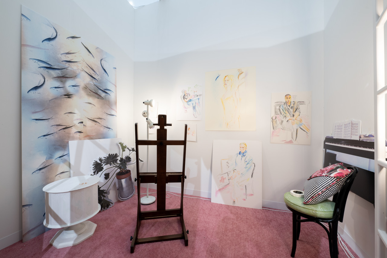 A color image of a room with white walls and pink carpet. A large painting depicting multiple fish leans on the left wall, along with a white canvas, and a cut-out of the picture of a potted plant. Across the room, a white table, a white lamp, a wooden easel and cushioned chair can be seen. Five colorful drawings of different sizes hang and lean on the back wall. A cut-out of a photograph of a piano with some sheet music hangs on the right wall.