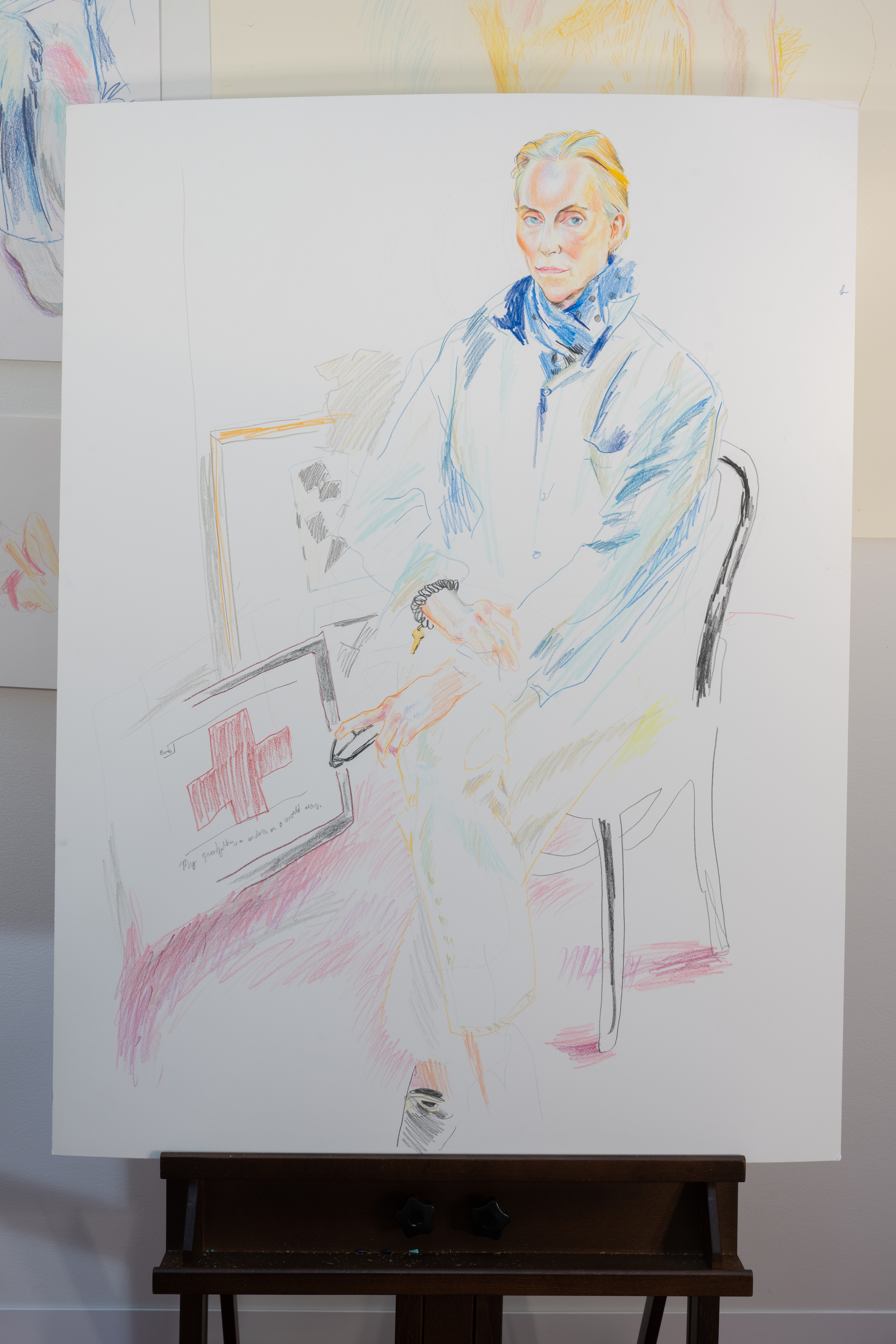A color image of an unframed colorful drawing reasting on a wooden easel. The drawing shows a figure elagantly sitting cross-legged on a black chair. In the background, two painting are on the floor laying against the wall.