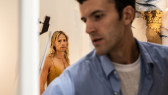 A color image of two figures. The figure in the foreground is blurry, and is looking towards the lower right corner. The figure in the background is focused and is looking straight at the viewer. There is a wooden column on the right.