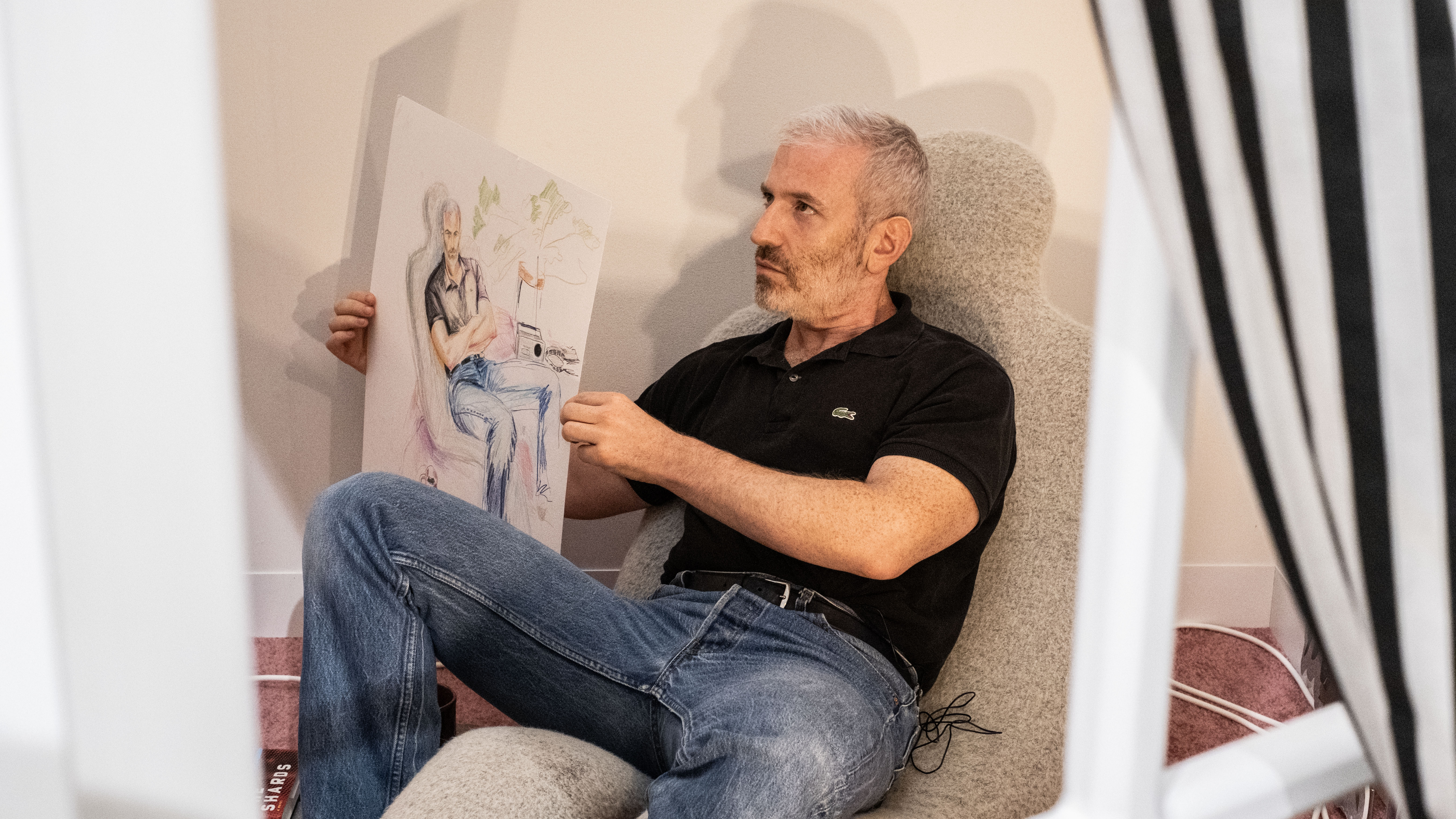 A color image of a man sitting down on a low sofa. He is seen, through a window, holding up a portrait of him that depicts him in this specific scenario.