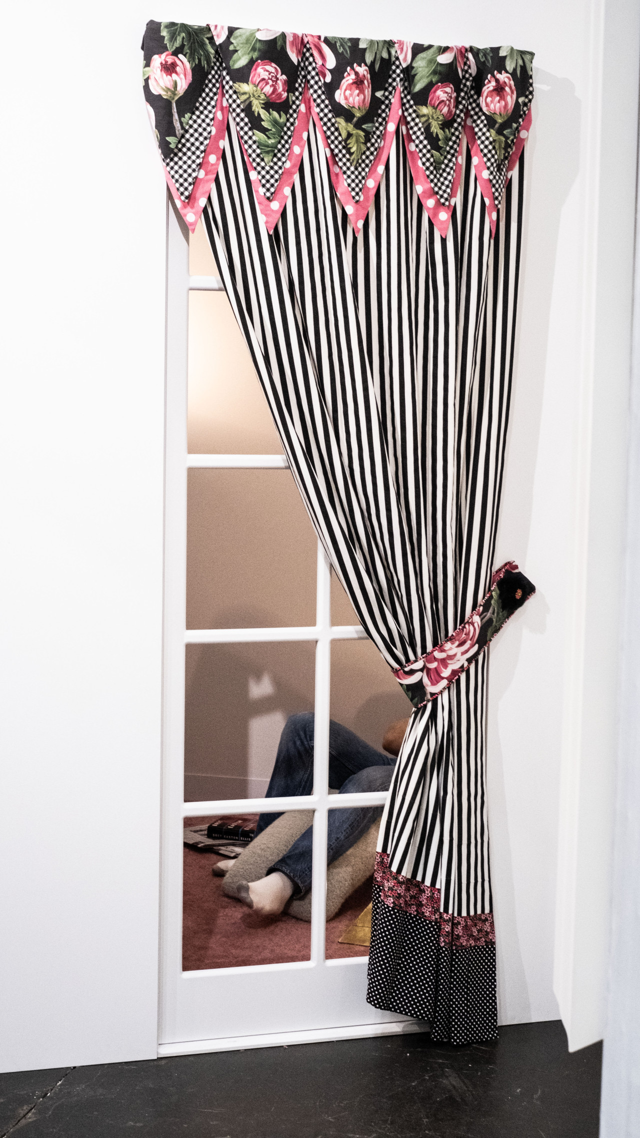 A color image of a white wall with windows and black and white curtains with pink flowers. Through the windows, a figure is seen laying down on a sofa. Only the legs are visible.