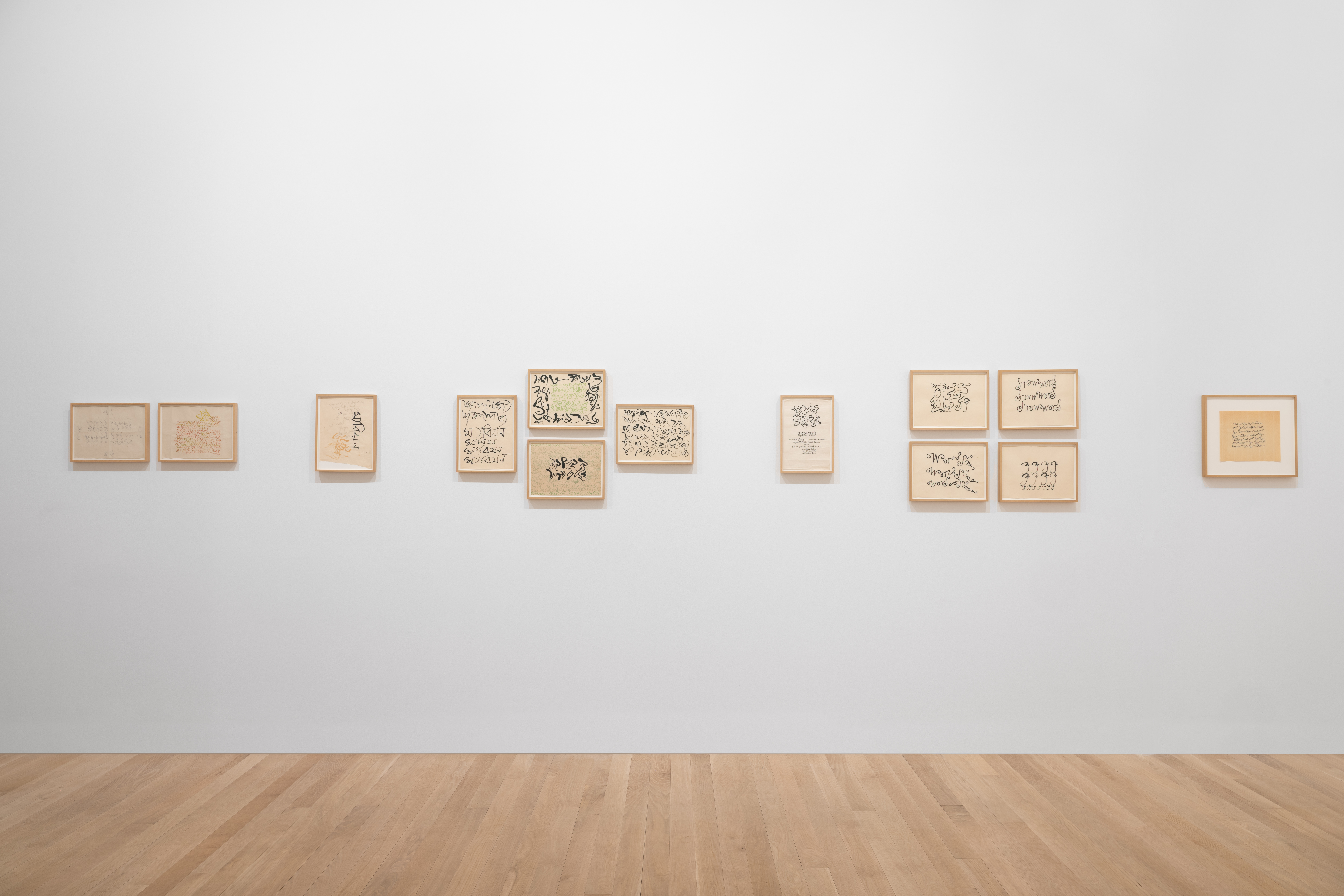 Alternating clusters and individual small framed works on paper hang on a white gallery wall. The works feature swooping, abstract calligraphic lines that resemble text.