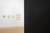 Installation view of white gallery wall featuring several small framed works on paper. Black wall featuring black text that reads 