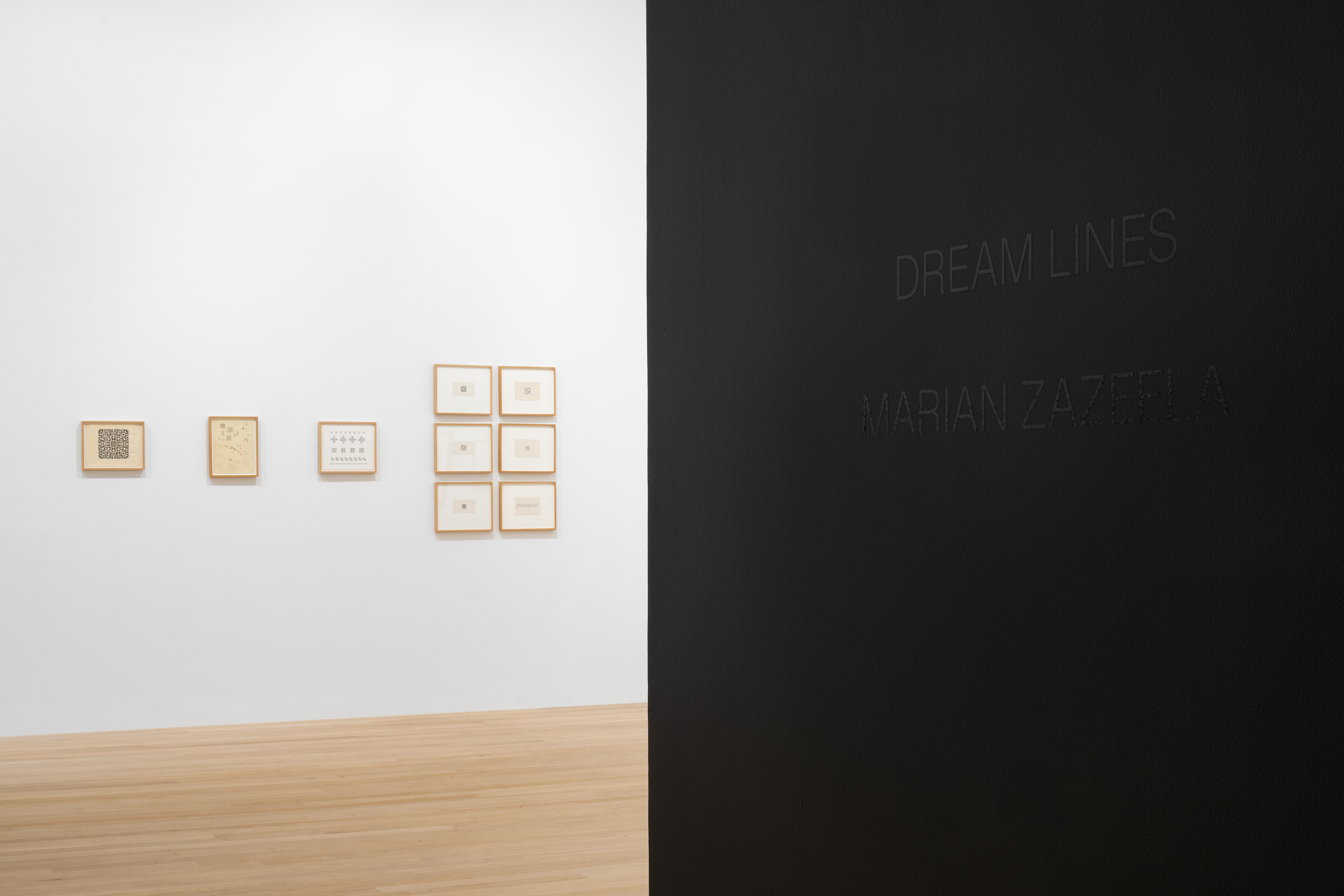 Installation view of white gallery wall featuring several small framed works on paper. Black wall featuring black text that reads 