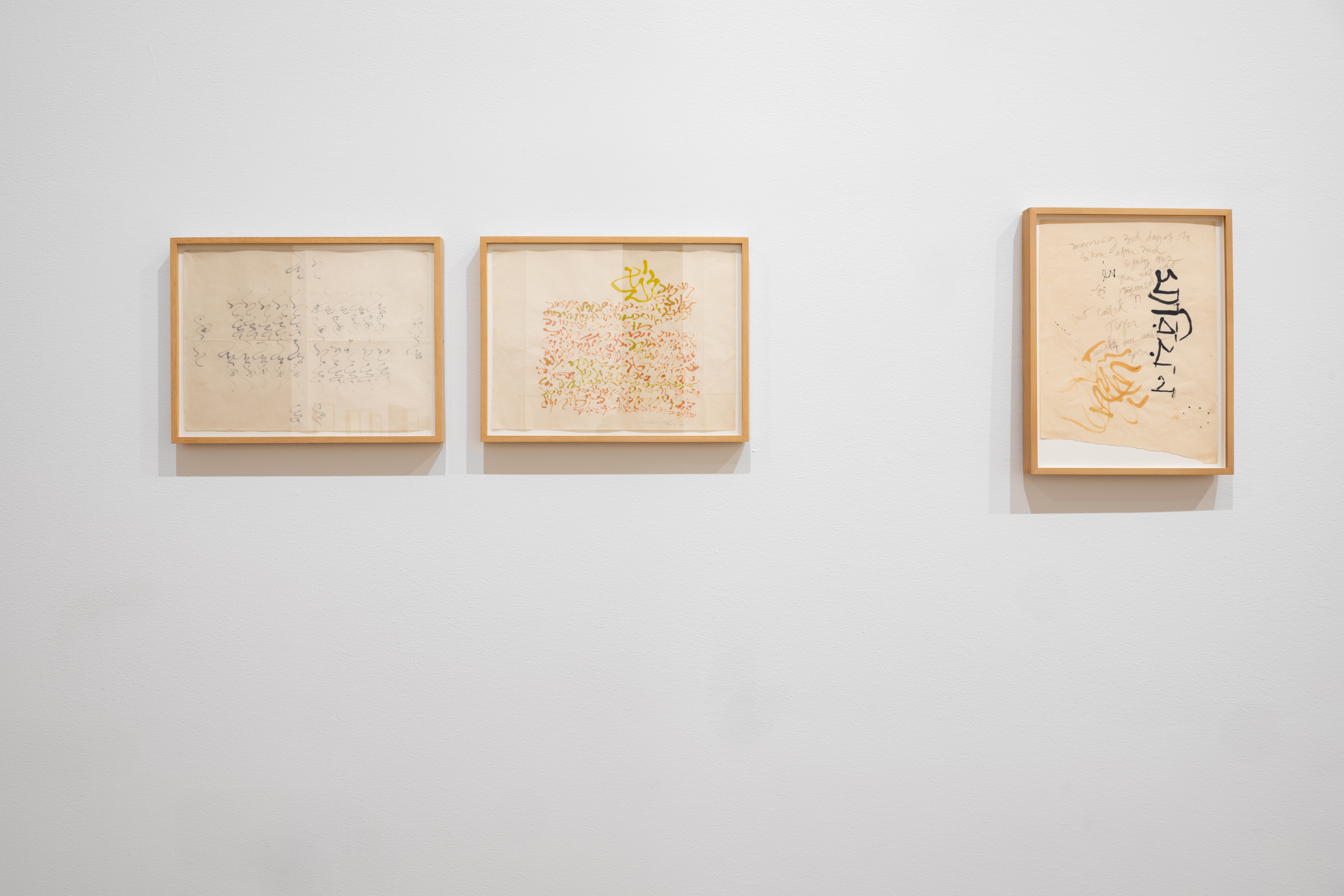 Closeup view of three small framed works on paper hanging on a white gallery wall. The works feature detailed, abstract calligraphic strokes in black, orange, and chartreuse.