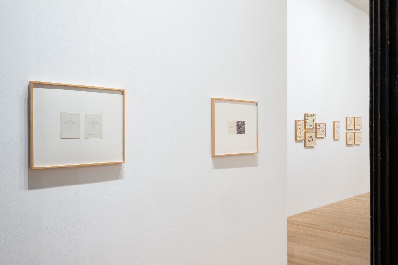 White gallery wall in foreground features two framed works on paper. The closer work features a diptych of delicately pencil-drawn crosses, while the further work is densely filled with calligraphic lines. Additional clusters of small framed works on paper hang on the wall in the background.