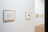 White gallery wall in foreground features two framed works on paper. The closer work features a diptych of delicately pencil-drawn crosses, while the further work is densely filled with calligraphic lines. Additional clusters of small framed works on paper hang on the wall in the background.
