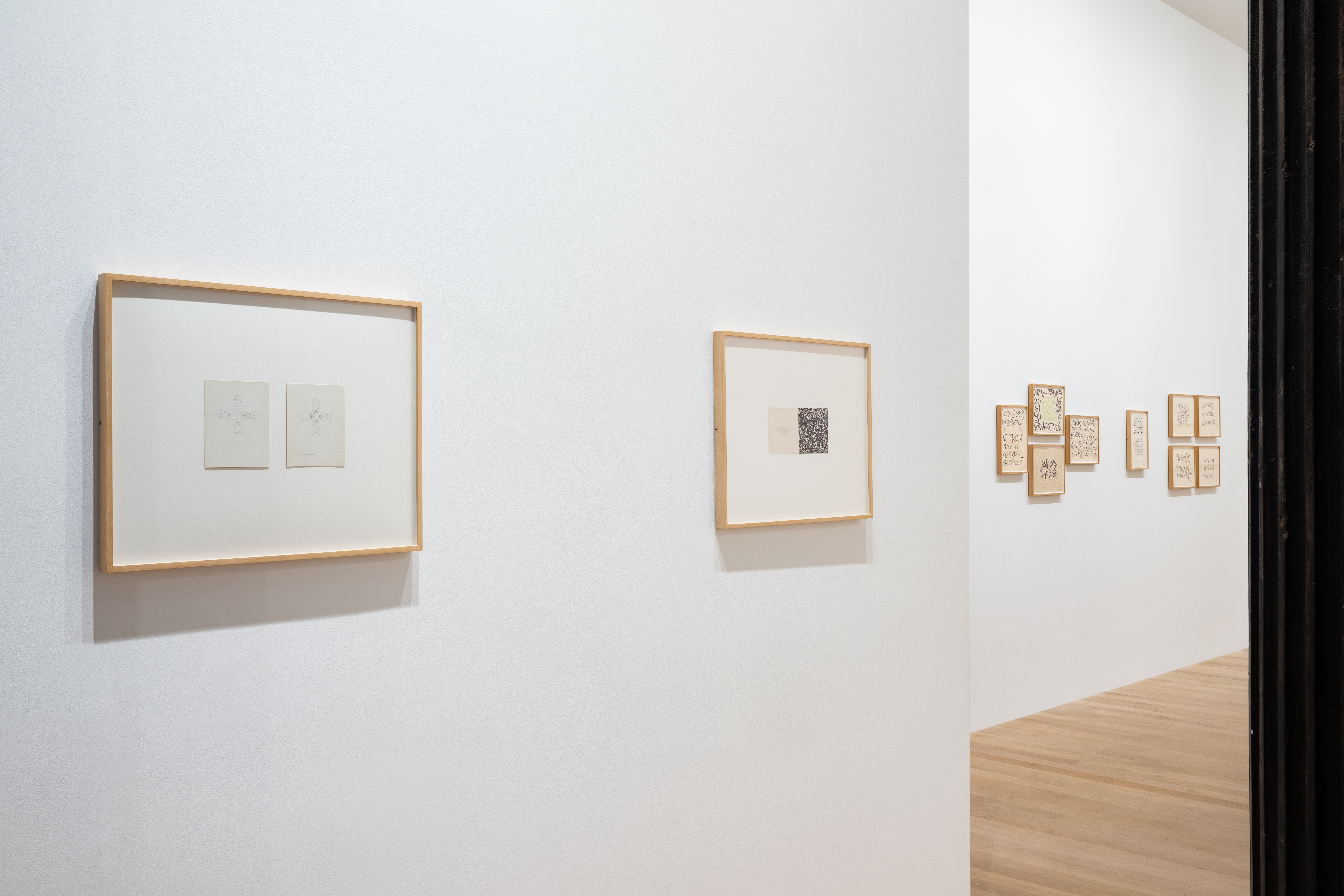 White gallery wall in foreground features two framed works on paper. The closer work features a diptych of delicately pencil-drawn crosses, while the further work is densely filled with calligraphic lines. Additional clusters of small framed works on paper hang on the wall in the background.