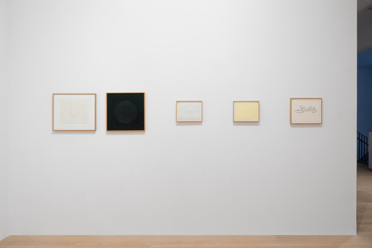 Framed works on paper hang on a white gallery wall. Some feature large circles composed of smaller ornamental swooping lines. Others feature abstract calligraphic strokes that resemble text.