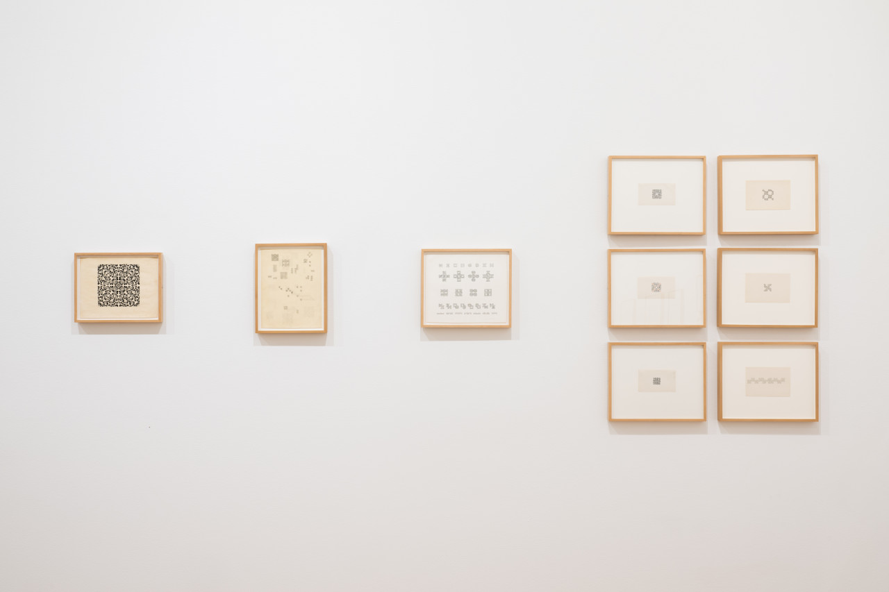 Various small drawings on paper in india ink and pencil hang on a white gallery wall. The works in pencil feature a looping ornamental symbol, drawn in various orientations within the individual box units of gridded paper to form larger structures.