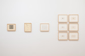 Various small drawings on paper in india ink and pencil hang on a white gallery wall. The works in pencil feature a looping ornamental symbol, drawn in various orientations within the individual box units of gridded paper to form larger structures.