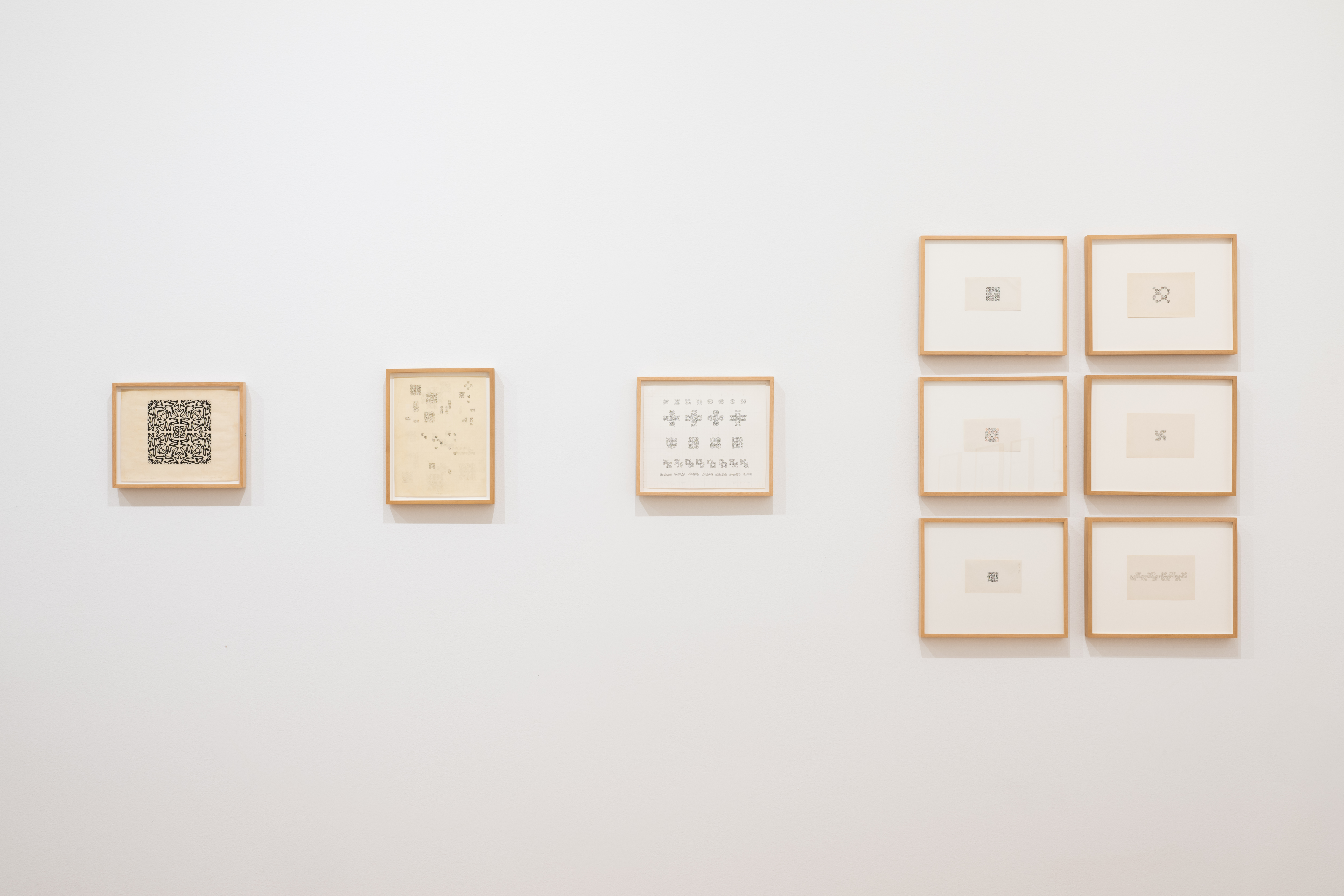 Various small drawings on paper in india ink and pencil hang on a white gallery wall. The works in pencil feature a looping ornamental symbol, drawn in various orientations within the individual box units of gridded paper to form larger structures.