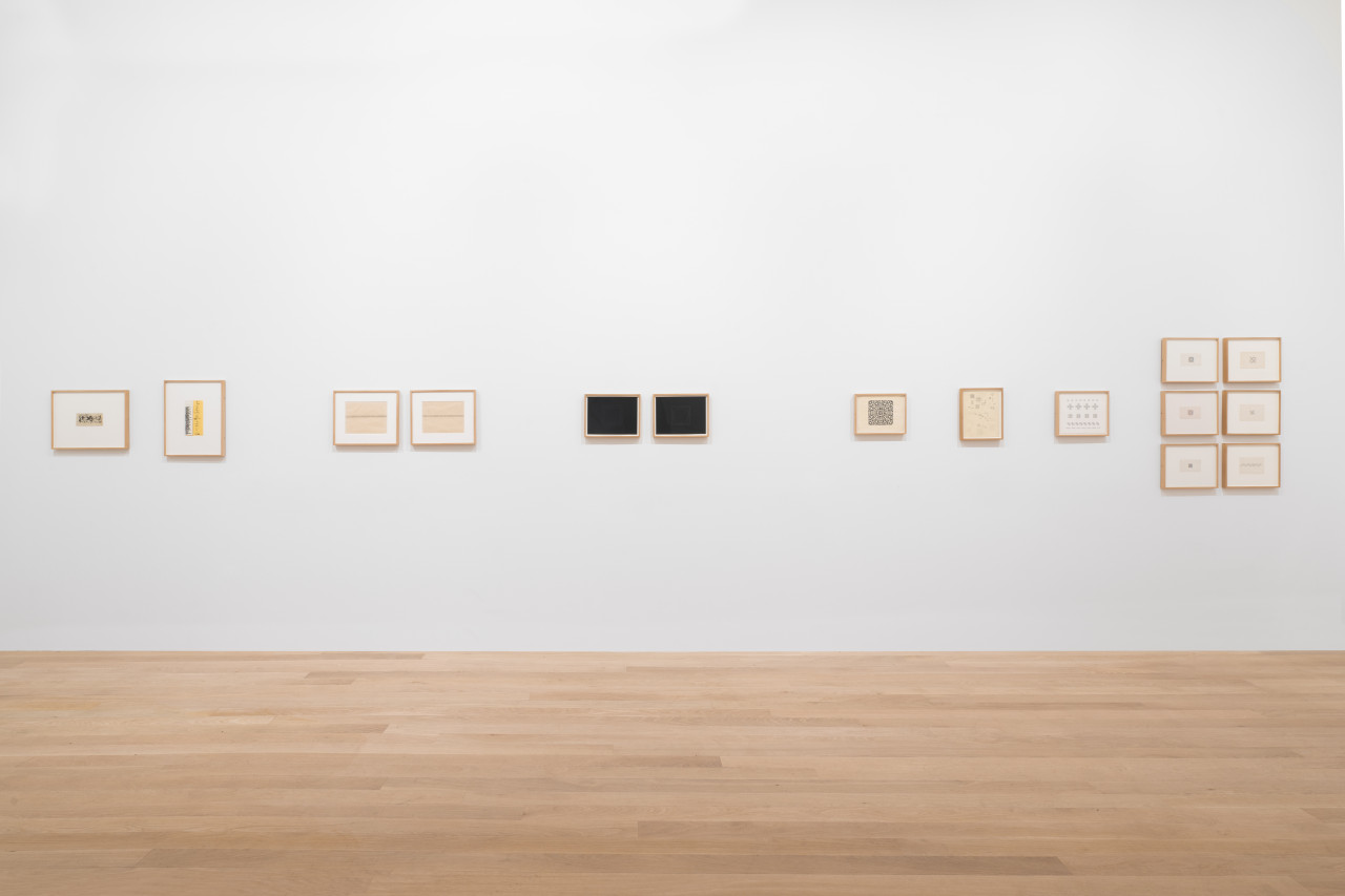 White gallery wall featuring various small framed works on paper, some featuring abstract ornamental bands, large concentric shapes against black ground, clusters of curled black lines organized into a square shape, and pencil-shaded boxes on gridded paper in various geometric shapes and configurations.