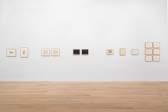 White gallery wall featuring various small framed works on paper, some featuring abstract ornamental bands, large concentric shapes against black ground, clusters of curled black lines organized into a square shape, and pencil-shaded boxes on gridded paper in various geometric shapes and configurations.