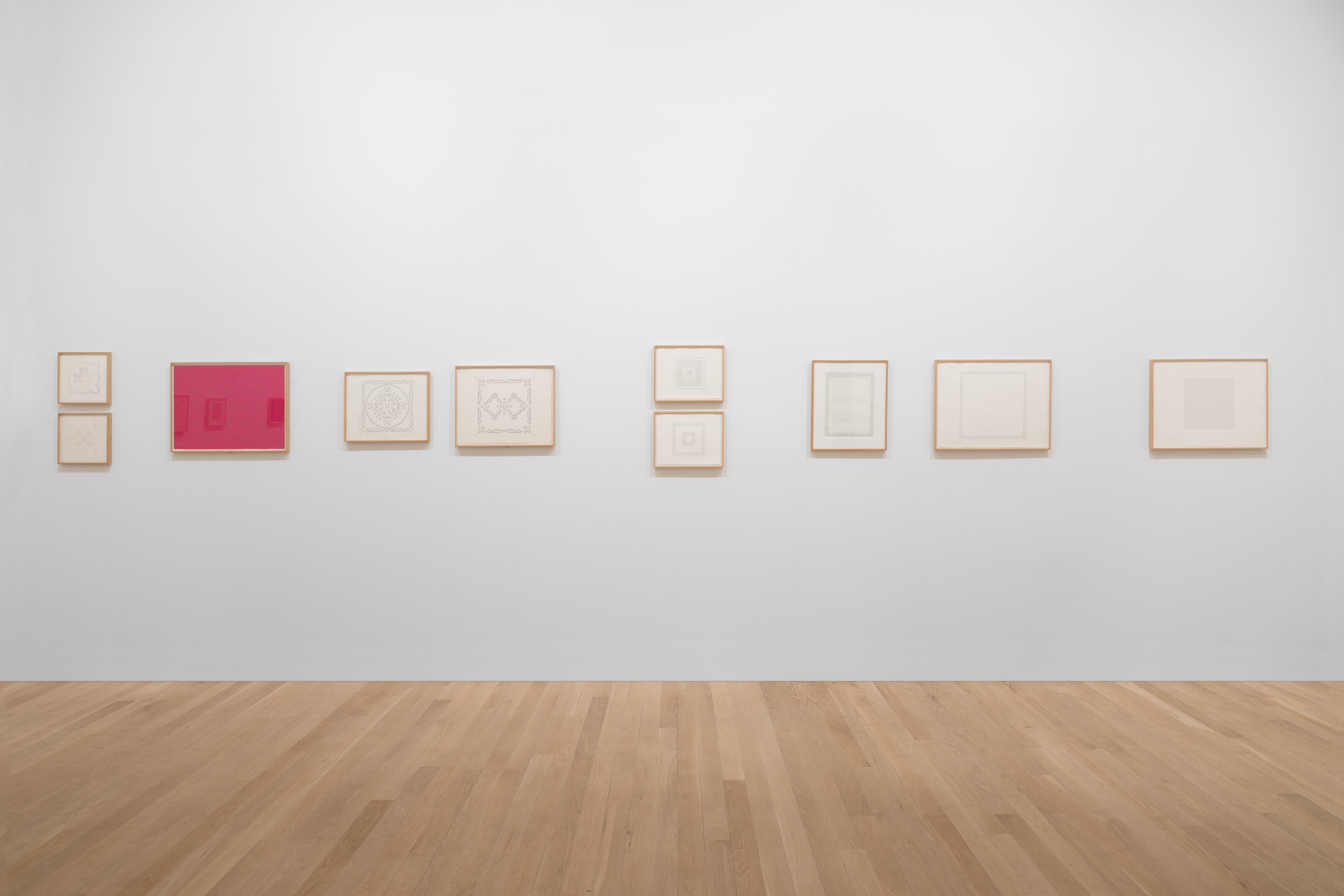 White gallery wall featuring various small framed works on paper, one on pink ground.
