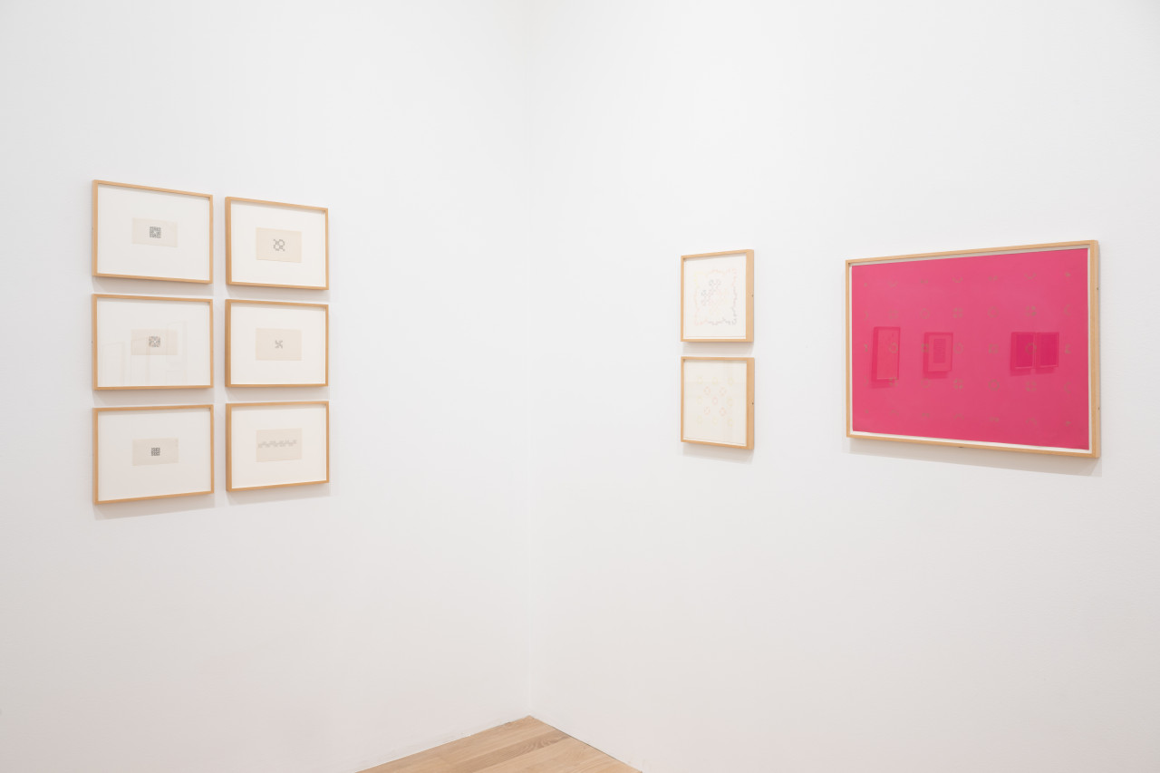 Various framed works on paper hang on two facing white gallery walls. Each features small looping ornamental symbols, drawn with pencil within the individual boxes of gridded paper, to form larger shapes and structures. The large work on the right wall features repeated ornamental symbols in gold on pink ground.
