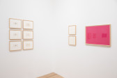 Various framed works on paper hang on two facing white gallery walls. Each features small looping ornamental symbols, drawn with pencil within the individual boxes of gridded paper, to form larger shapes and structures. The large work on the right wall features repeated ornamental symbols in gold on pink ground.