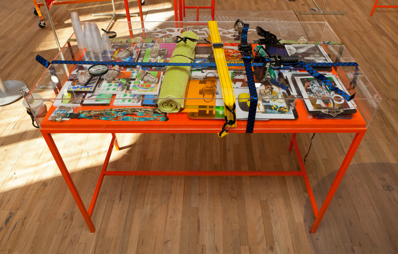 A glass case filled with colorful paraphernalia sits on an orange table. The case is bound lengthwise and widthwise with straps. On top of the case sit two magnifying glasses and several stacks of plastic cups.