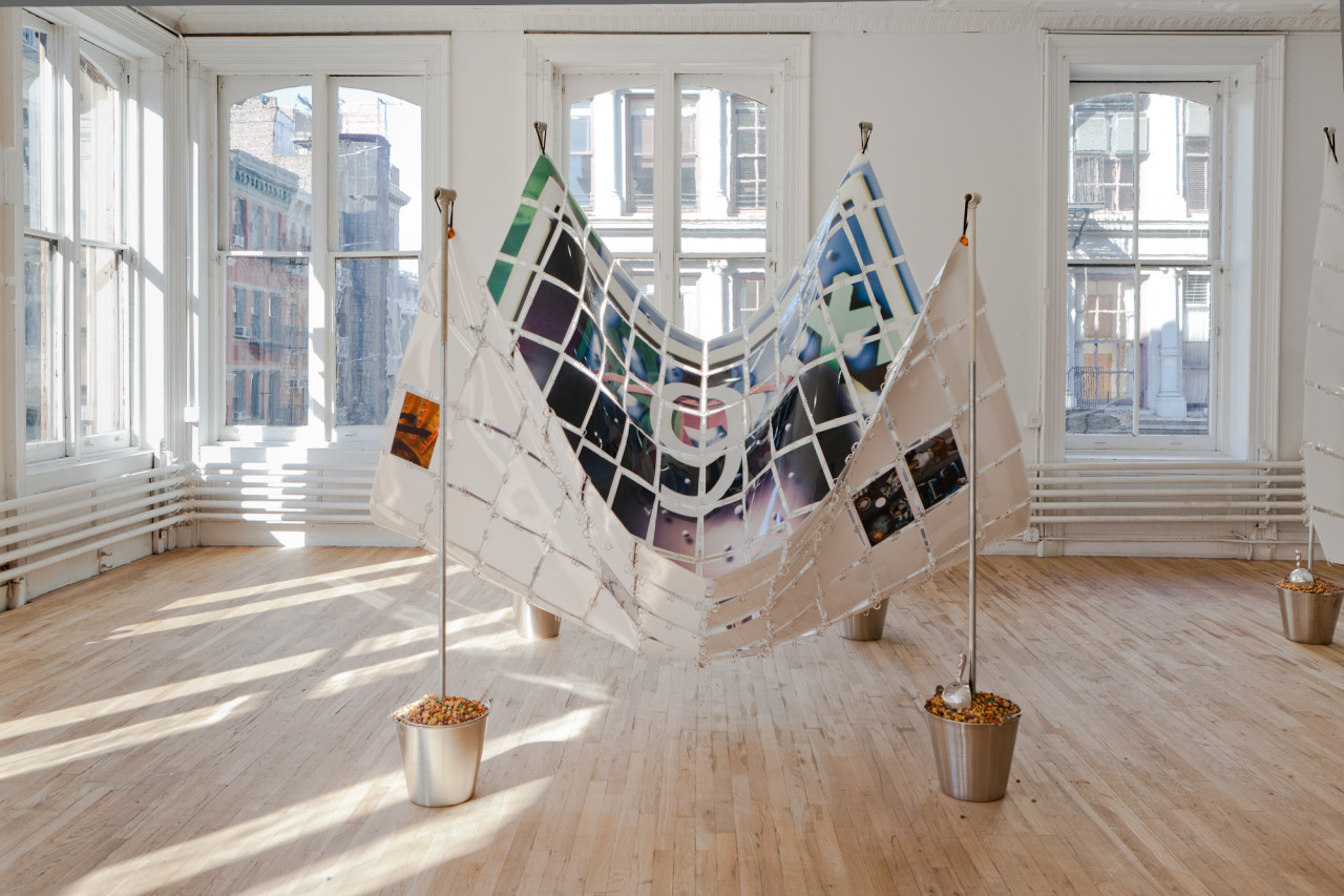 An image of a large number "9" printed across a series of rectangular pieces of paper that are linked together by metal rings such that they form a large, draping sheet. The sheet of linked papers is held in the air by four poles supporting each of its four corners. The poles are mounted in silver buckets filled with multicolored objects.