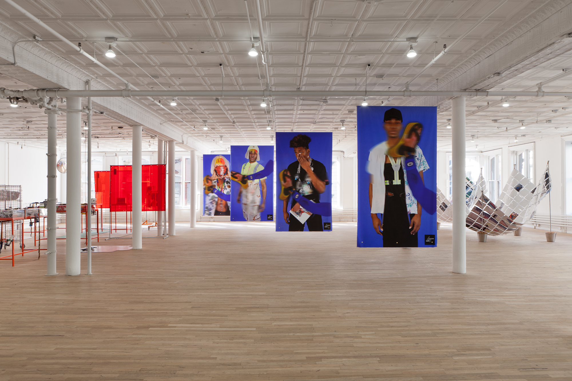 A wide view of an installation in a large exhibition space. Four images of figures posing in various outfits hang from the ceiling in diagonally decreasing proximity. To the right of the space, an abstract sculptural piece is displayed. To the left are several orange tables and two orange, transparent rectangles supported by metal frames.