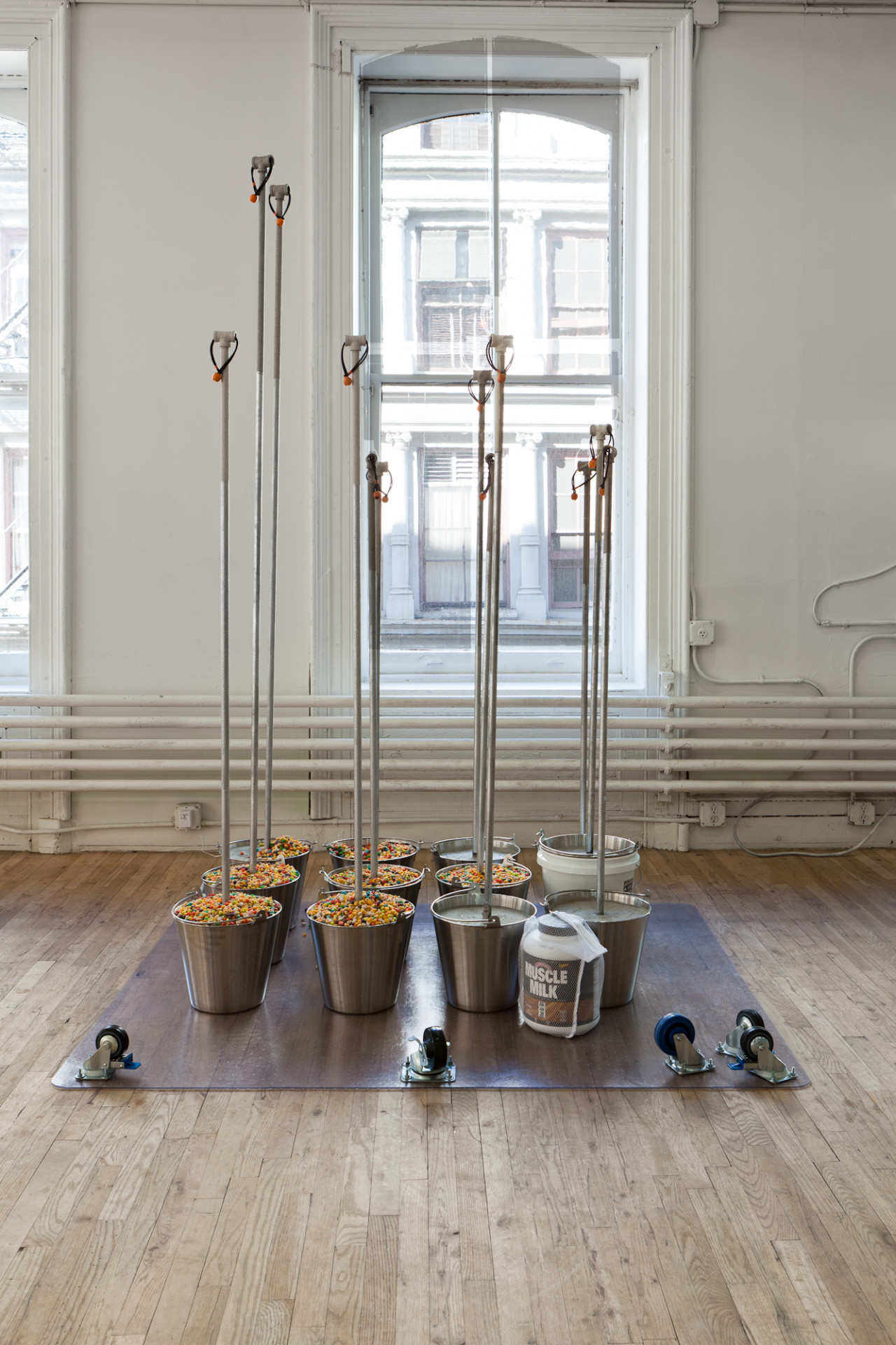 A plastic surface with five wheels lies upturned on a wooden floor. Eleven metal buckets sit on the upturned surface, some filled with multicolored objects. A silver pole extends out from each of the buckets. In front of the buckets sits a large container of Muscle Milk enclosed in a net bag.