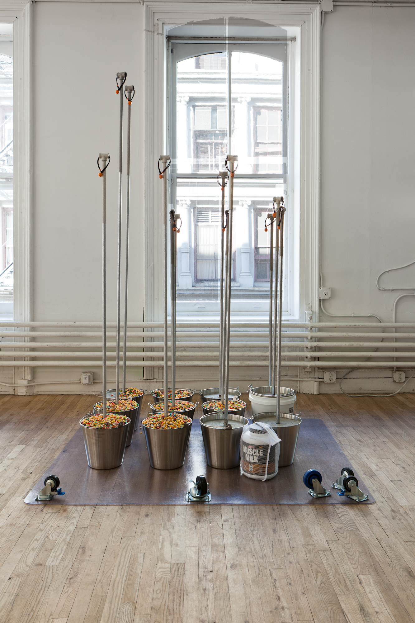 A plastic surface with five wheels lies upturned on a wooden floor. Eleven metal buckets sit on the upturned surface, some filled with multicolored objects. A silver pole extends out from each of the buckets. In front of the buckets sits a large container of Muscle Milk enclosed in a net bag.