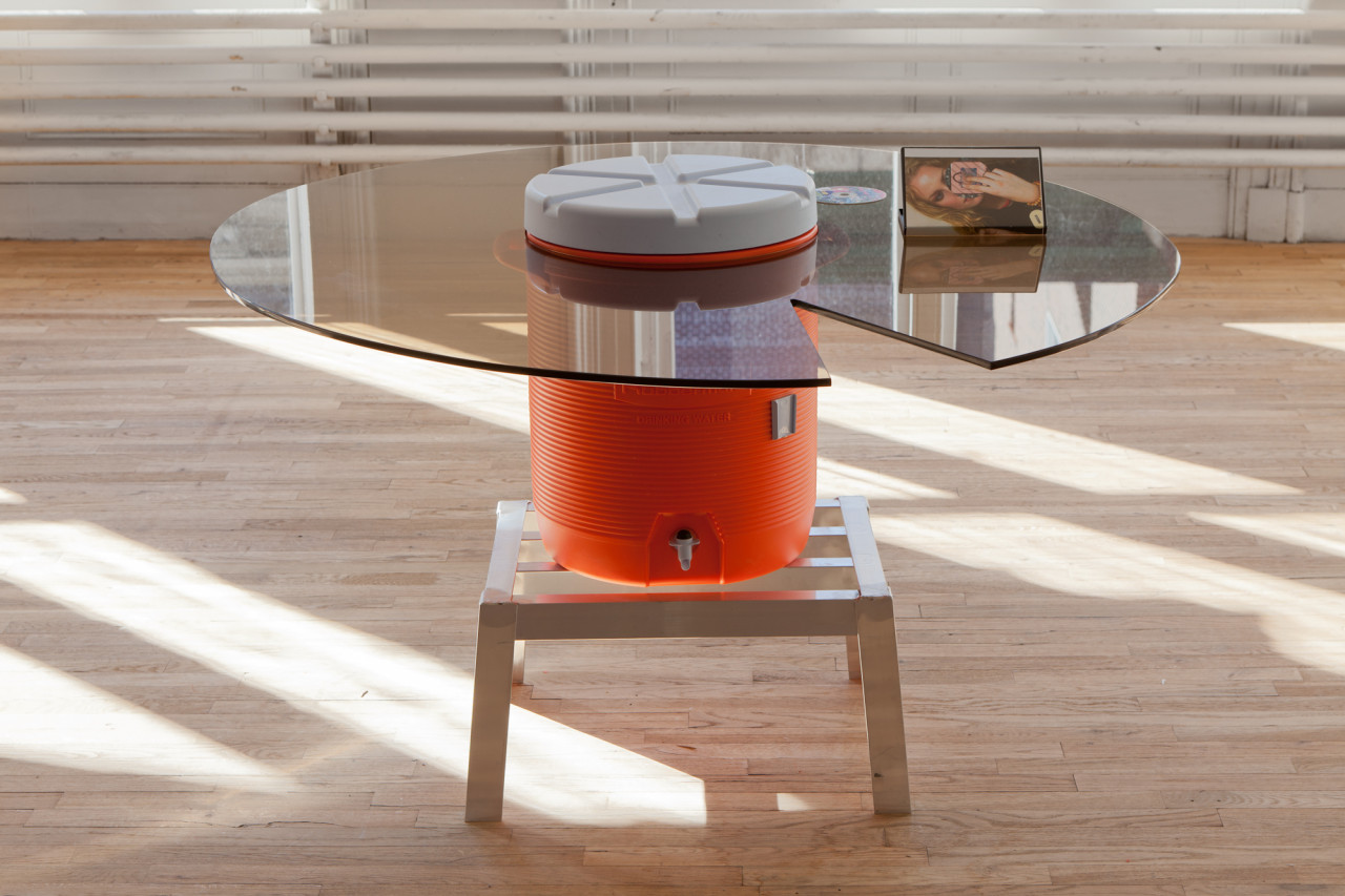An orange water dispenser sits on a low metal stool in a gallery space. Encircling the top portion of the dispenser is a round glass table, with a wedge-shaped cut-out removed from its right side. A photo of a woman sits on top of the glass surface.